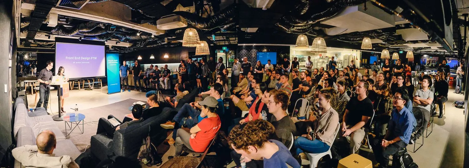 A panoramic shot of a packed SydCSS meetup — two presenters on stage in front of a large screen, with rows of engaged attendees filling the room.